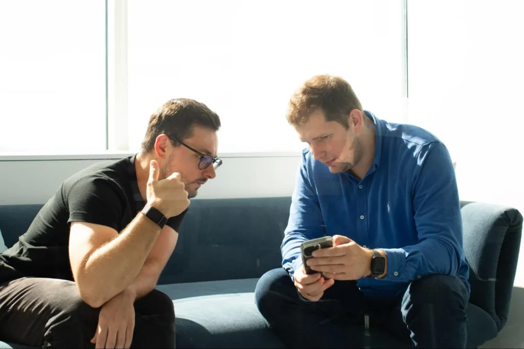 Two specialists from Black Label company sitting on a couch, with one of them showing something on his smartphone to the other, who is looking intently at the screen.
